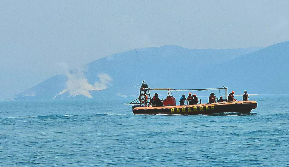 Perahu karet Basarnas berisi petugas penyelamat berpelampung oranye sedang mengarungi laut Selat Sunda dengan latar belakang kepulan asap putih Gunung Anak Krakatau di Kabupaten Pandeglang Banten.