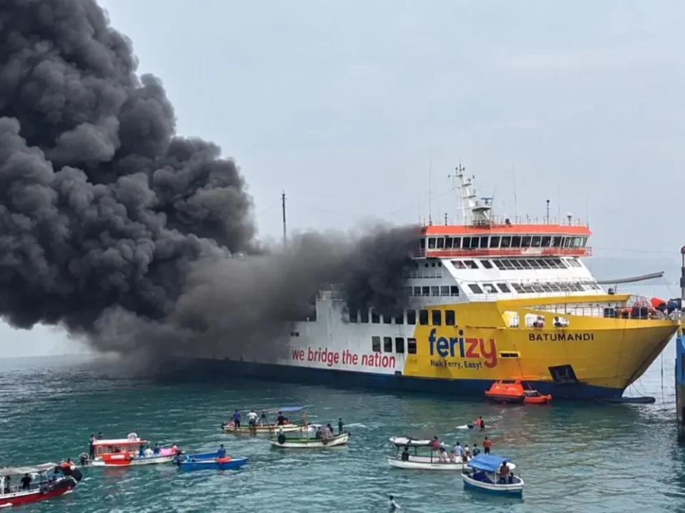 Foto horizontal dari jarak dekat menunjukkan Kapal Motor Penumpang (KMP) Batumandi berwarna kuning dan putih dengan logo "ferizy" sedang mengeluarkan asap hitam tebal yang masif dari bagian belakang. Di sekitar kapal laut yang sedang terapung di perairan jernih tersebut, tampak sekitar enam perahu motor kayu kecil milik nelayan dan satu sekoci penyelamat oranye merapat untuk mengevakuasi para penumpang.